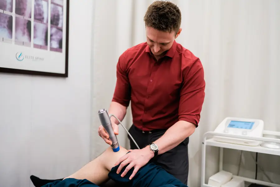 Clinician pressing a radial shockwave applicator against a patient's knee, with the therapy console on a trolley beside the treatment table.